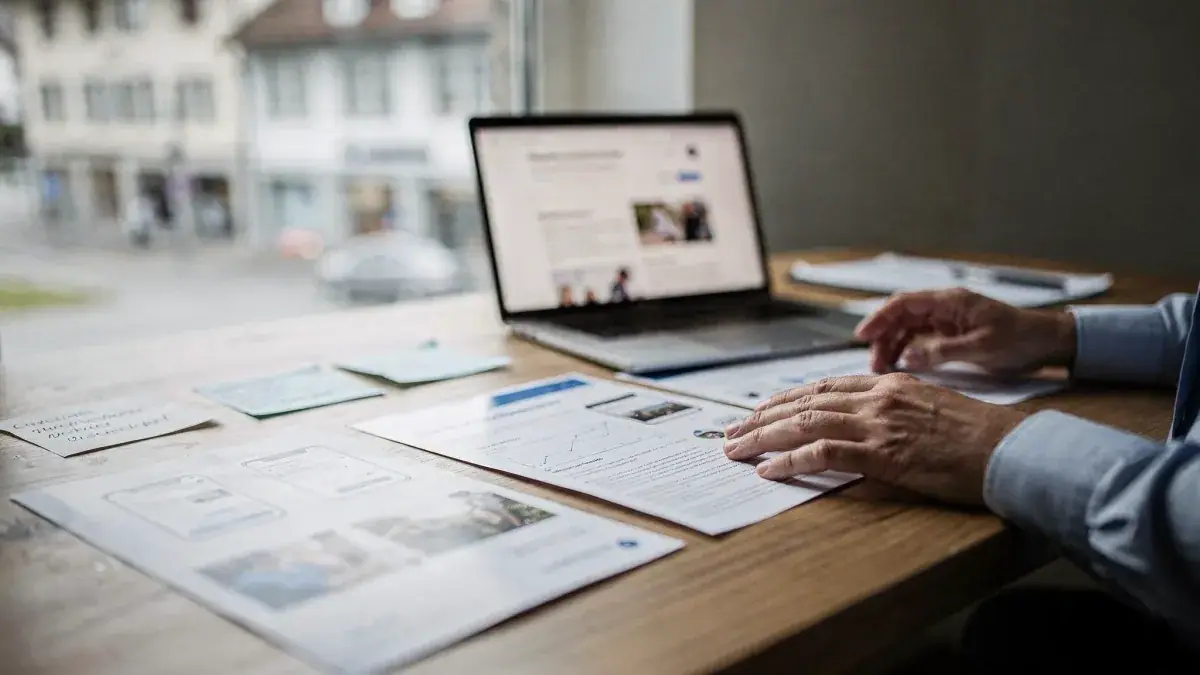 Person am Laptop mit Website und ausgedruckten Layouts am Fenster